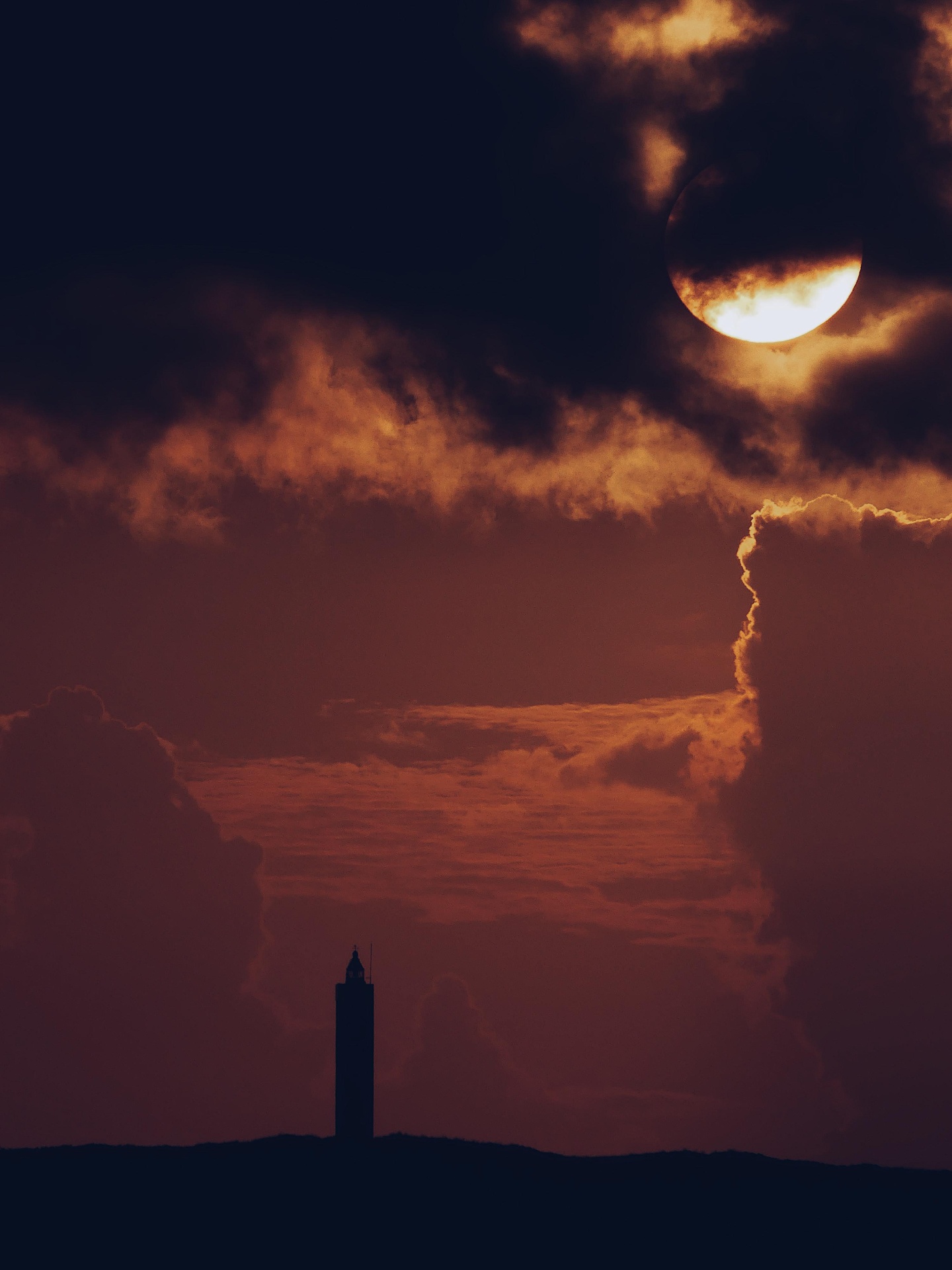 Sun breaking through clouds above a distant lighthouse at sunset.