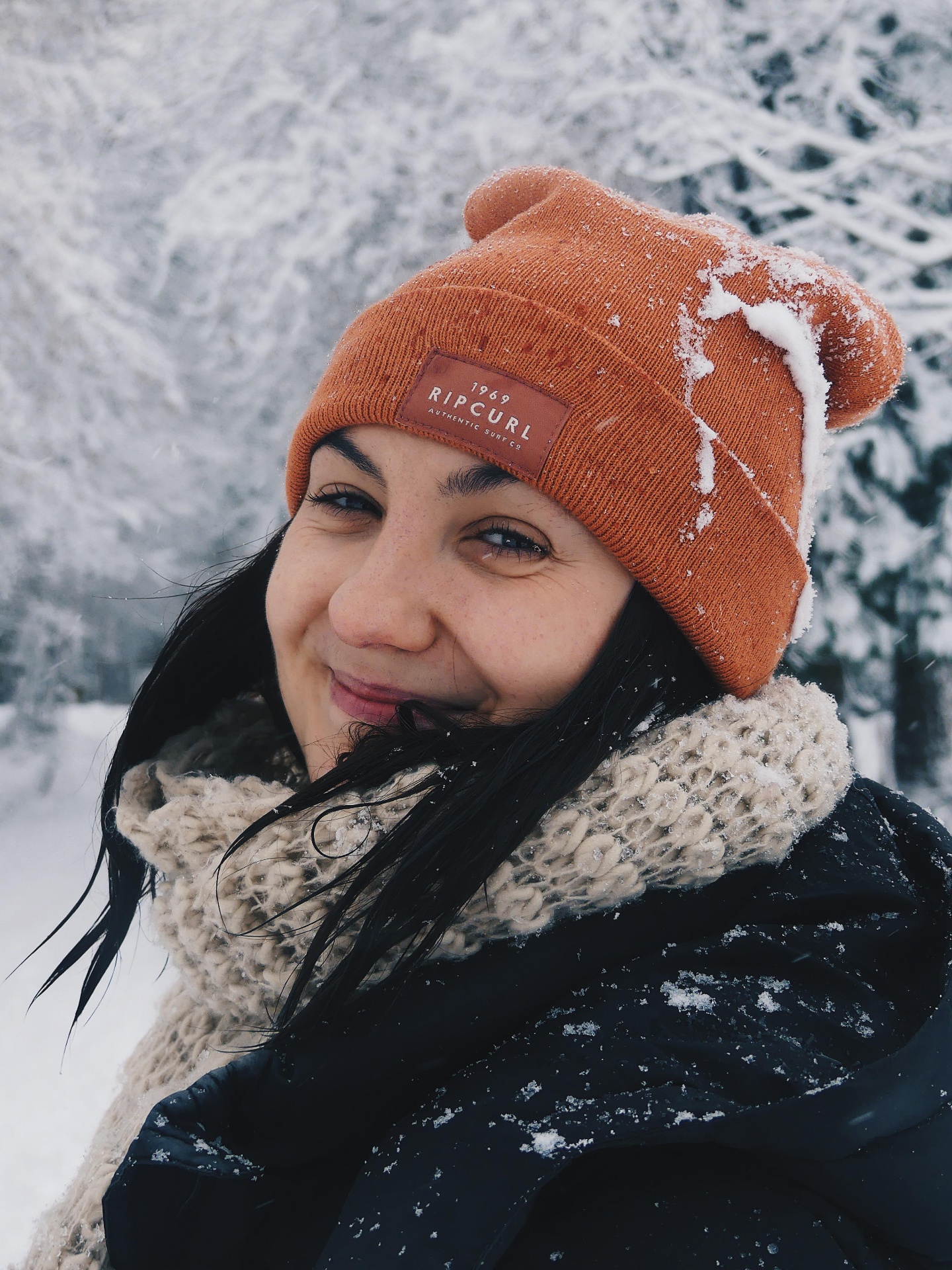 Smiling portrait in snowfall with an orange beanie and knitted scarf.