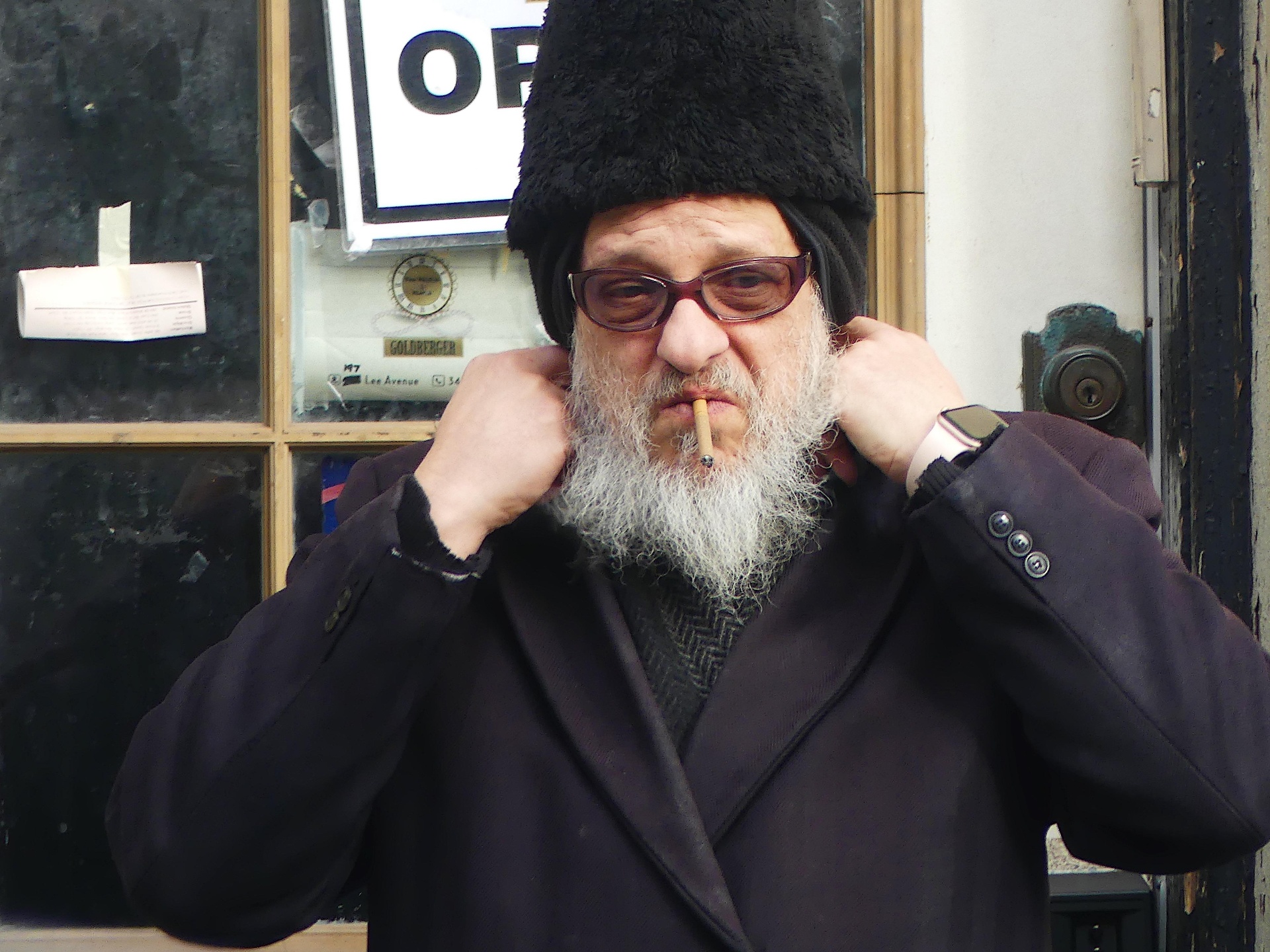 Portrait of an older man with glasses, beard, and cigarette outside a storefront.