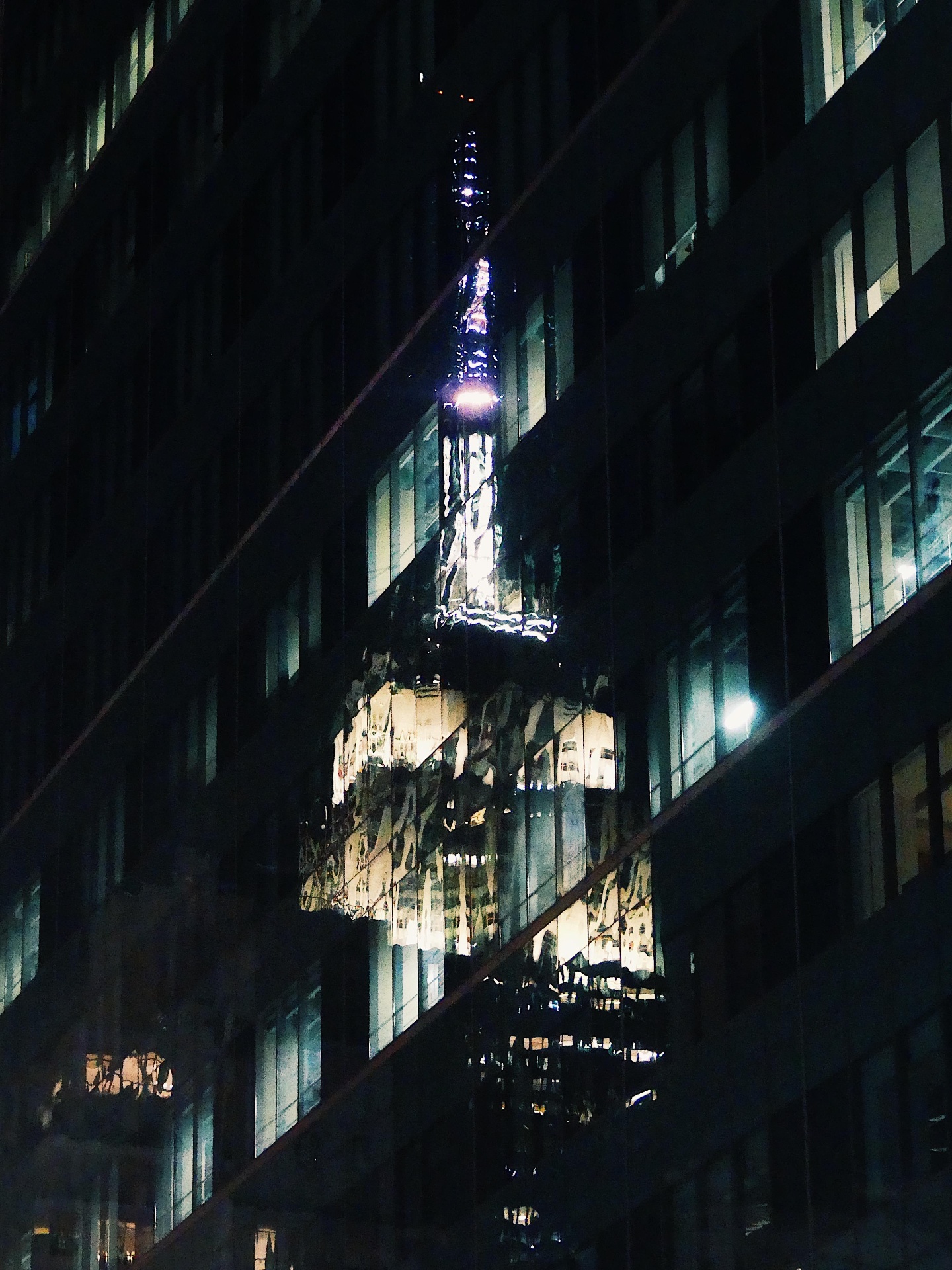 Spire of a skyscraper reflected in the glass of a neighbouring building at night.