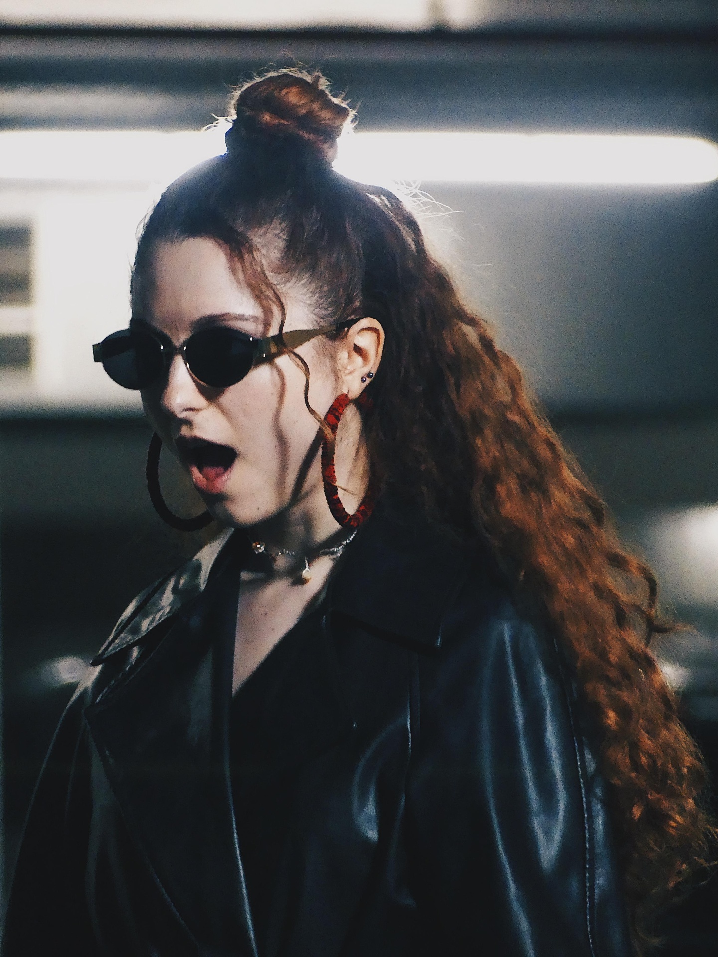 Red-haired woman in sunglasses and leather jacket under bright fluorescent light.