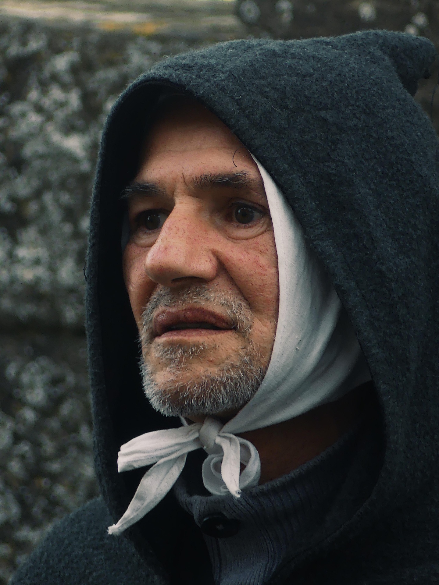 Weathered portrait of a hooded man with a white wrap against stone.